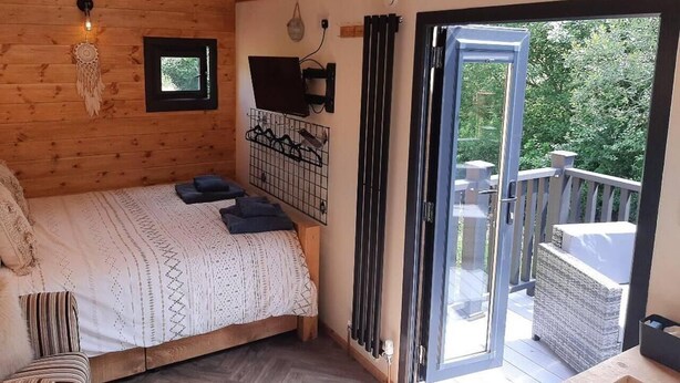 A wooden cabin bedroom with a bed, a small window, a wall-mounted TV, and a balcony with a wicker chair. - Min yr Afon - Cabin - Sleeps 2 - Hot Tub - Parking (Anglesey)
