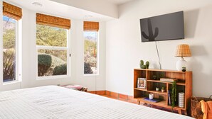 A bedroom with a large bed, two windows with blinds, a wooden bookshelf, and a wall-mounted TV. - Foothills Southwest Sanctuary w/ Pool & Views (Tucson)