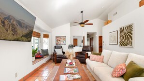 A spacious living room with a large flat-screen TV, a ceiling fan, a coffee table with magazines, a sofa with patterned cushions, and a large window with a mountain view. - Foothills Southwest Sanctuary w/ Pool & Views (Tucson)