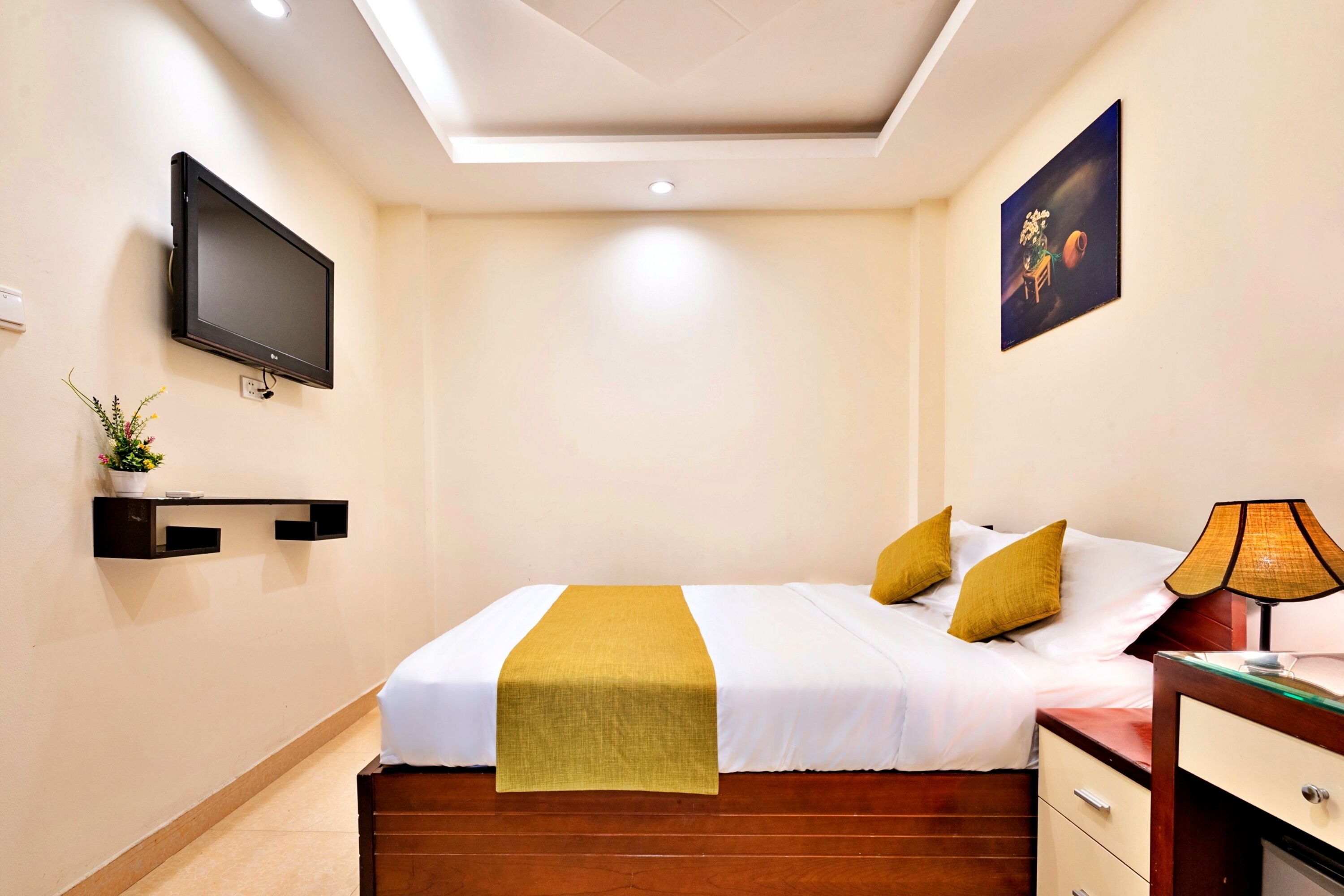 A hotel room with a bed, bedside table, lamp, TV, and a painting on the wall.