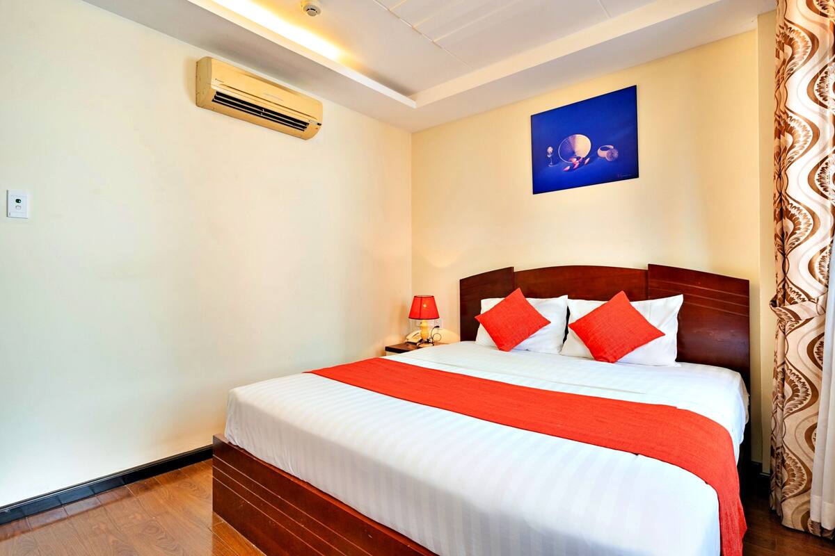 A neatly made bed with white and red bedding, a red throw, and a wooden headboard. There is a nightstand with a red lamp, a wall-mounted air conditioner, and a framed picture above the bed.