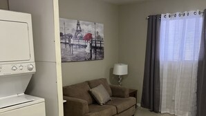 Una sala de estar con un sofá beige, un electrodoméstico blanco, un cuadro de una pareja y una ventana con cortinas.