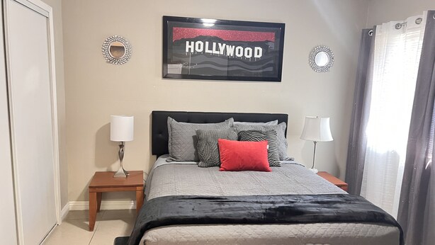 Un dormitorio con cama, dos mesitas de noche, un espejo, un cuadro en la pared y una ventana con cortinas.