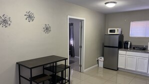Una cocina equipada con refrigerador, microondas y cafetera. Hay una mesita con dos repisas. Las paredes son lisas y el piso, de baldosas.