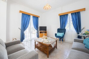 Un salon moderne avec une table basse en bois, des canapés gris, un fauteuil bleu et un téléviseur posé sur un meuble en bois.