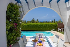 Uma área de refeições ao ar livre com vista para a piscina, cadeiras de vime branco e uma mesa posta com comida e bebidas.