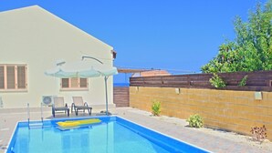 A swimming pool with lounge chairs and umbrellas, a wooden fence, and a view of the sea. - Villa Majestic in Agia Marina Chrysochous (Ayia Marina Chrysochous)