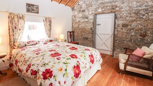 Une chambre avec un plafond en bois, un mur en pierre, un lit avec une literie à motifs floraux, une chaise et une porte.