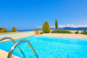 A clear blue pool with a metal ladder, surrounded by neatly trimmed bushes and a view of the ocean and distant mountains. - Villa Styliani in Fasli (Droushia)