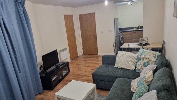 A living room with a sofa, a small white table, a TV, and a kitchen area in the background. - Central 2-bedroom Apartment, Chelmsford, Parking (Chelmsford)