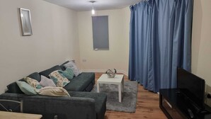 A living room with a dark green sofa, a small white table, a TV, and blue curtains. - Central 2-bedroom Apartment, Chelmsford, Parking (Chelmsford)