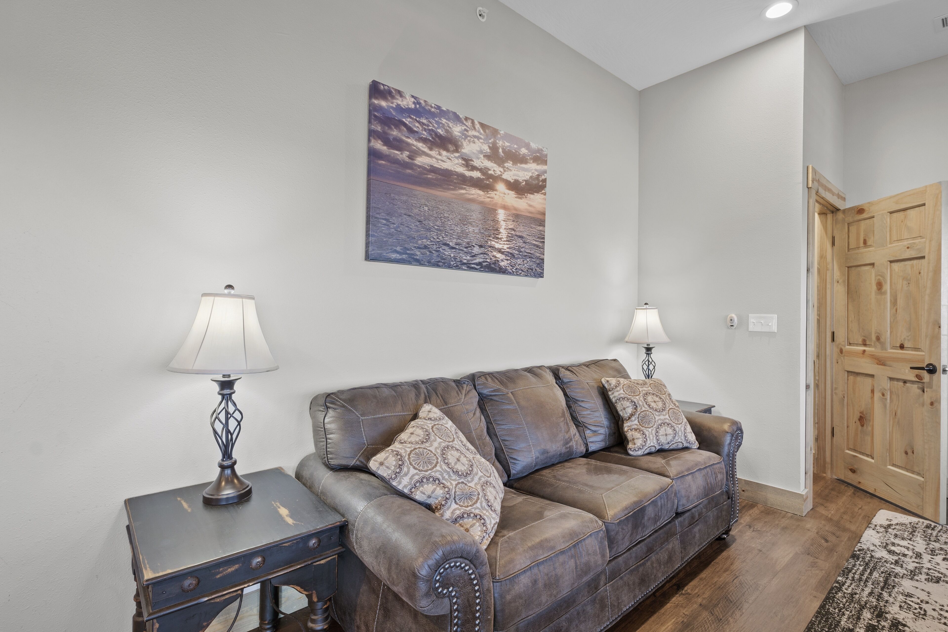 A living room with a brown leather sofa, two patterned throw pillows, a side table with a lamp, and a painting of a sunset on the wall.