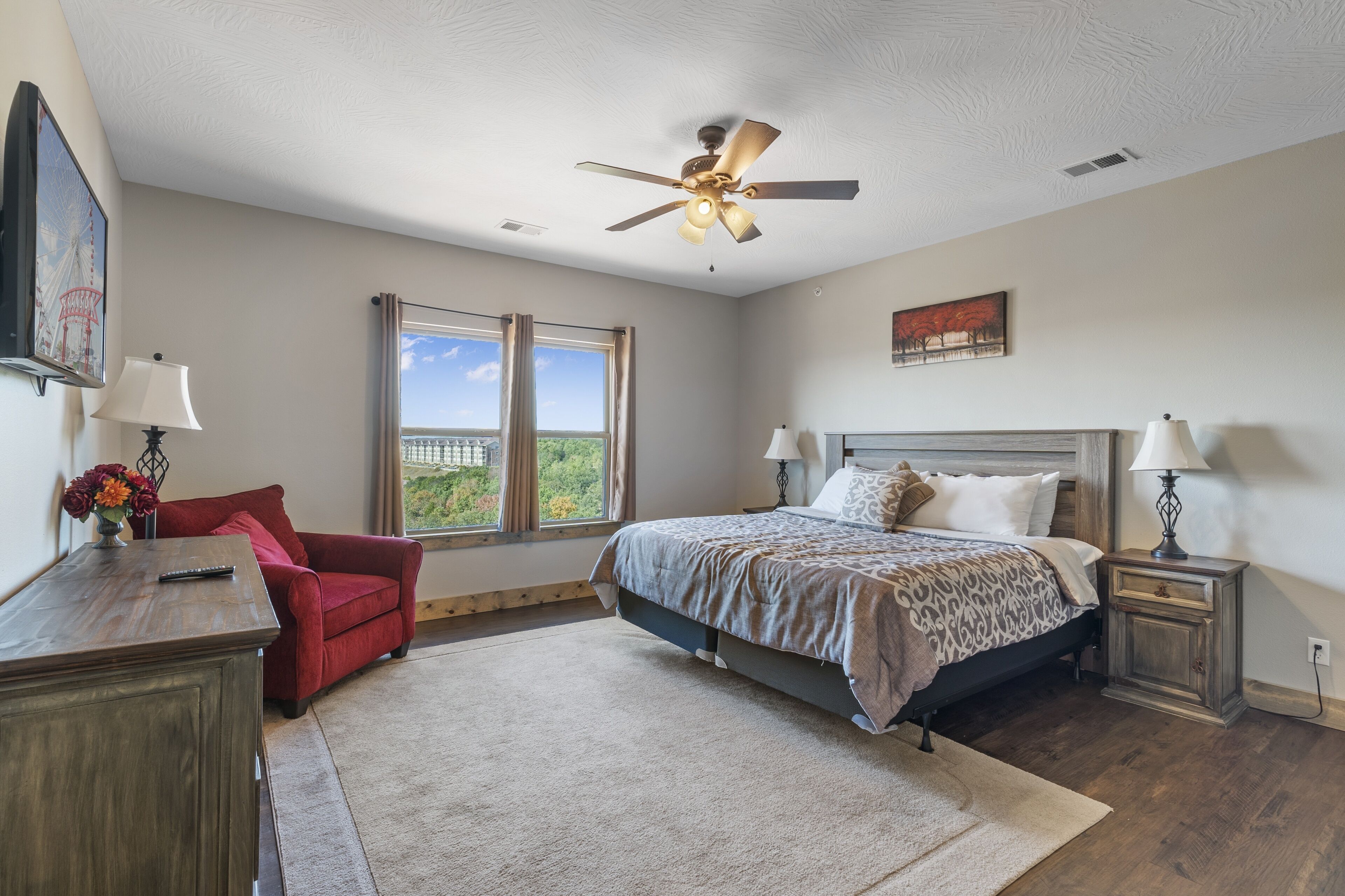 A bedroom with a bed, a red chair, a nightstand, a ceiling fan, and a window with a view.