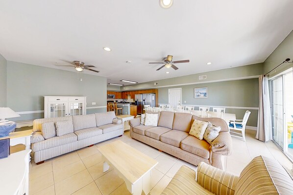 A spacious living area with two sofas, a coffee table, ceiling fans, and a kitchen in the background.