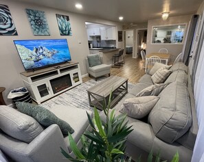 A modern living room with a grey sofa, a coffee table, and a TV on a wooden stand. There is a kitchen in the background with white cabinets and a dining area with a white table and chairs. - Cheerful Two Bedroom Bungalow Newly Remodeled! Arcade games, Air Hockey Table. (Eureka)