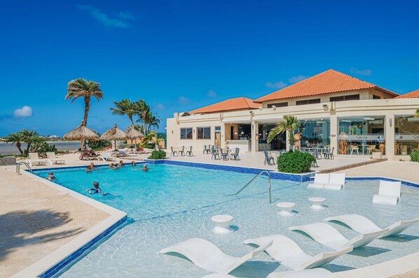 Zona de piscina con sillas de descanso, palmeras y un edificio con tejado de azulejos.
