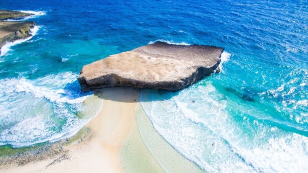 Ein Strand mit einer großen Felsformation, umgeben von klarem, blauem Wasser und einem sandigen Ufer.