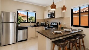Eine moderne Küche mit einem Edelstahlkühlschrank, einer Mikrowelle, einem Backofen und einer Kaffeemaschine. Zwei Pendelleuchten, ein Fenster mit Blick auf Palmen und eine Bar mit Hockern.