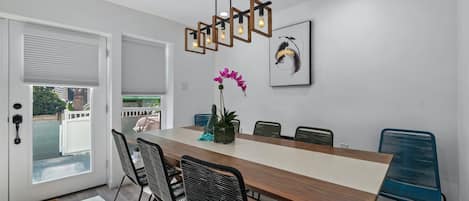 Un coin repas avec une table en bois, des chaises noires, un luminaire suspendu moderne et une vue à travers une porte vitrée.