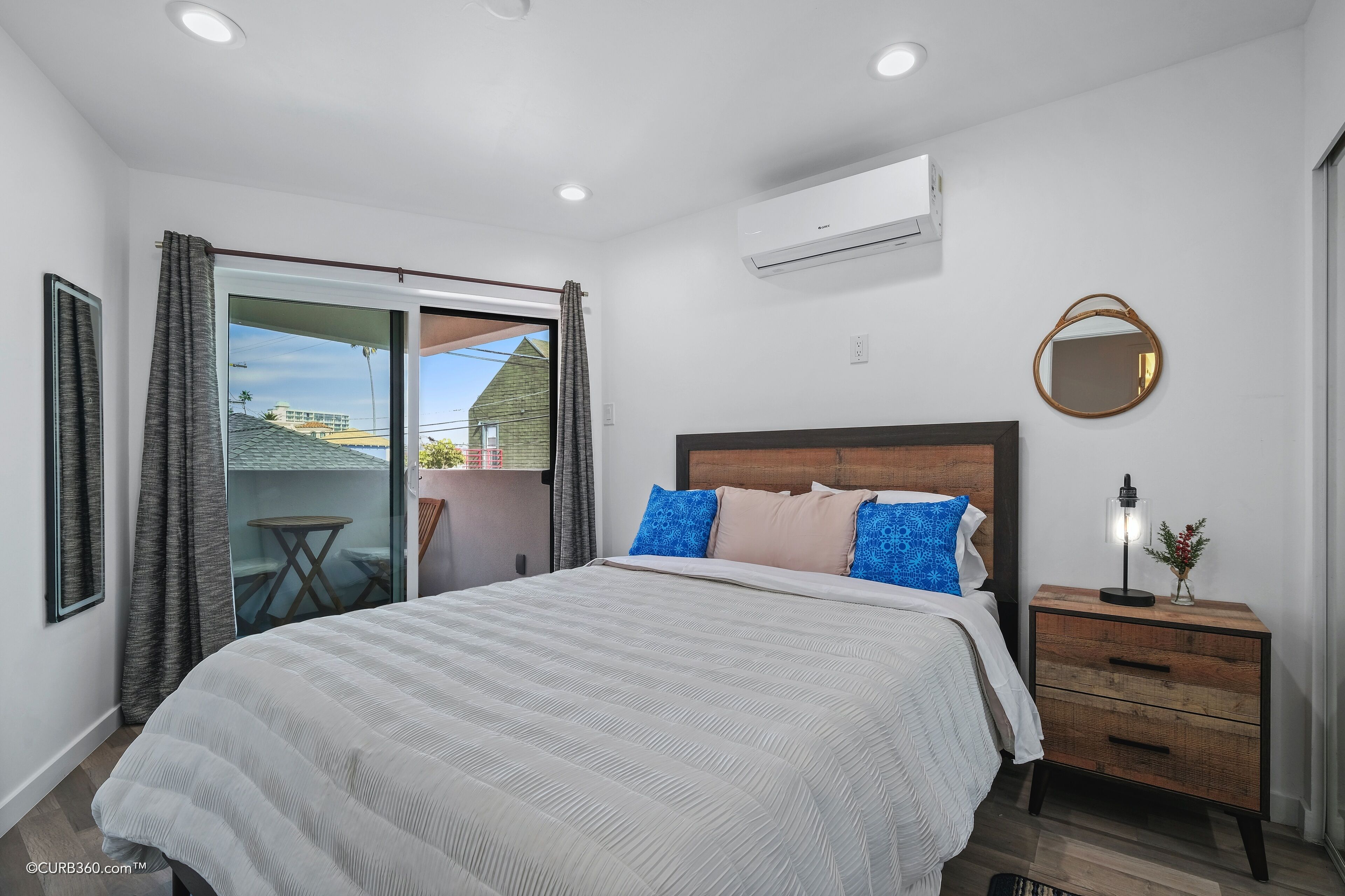 A bedroom with a large bed, a wooden nightstand, a round mirror, and a view of a balcony with a table and chairs.