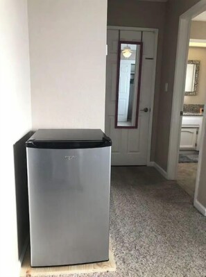 Compact refrigerator in a hallway with a carpeted floor, adjacent to a door with a mirror above.