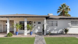 A single-story house with a covered porch, a front door, and a small garden with palm trees and shrubs.
