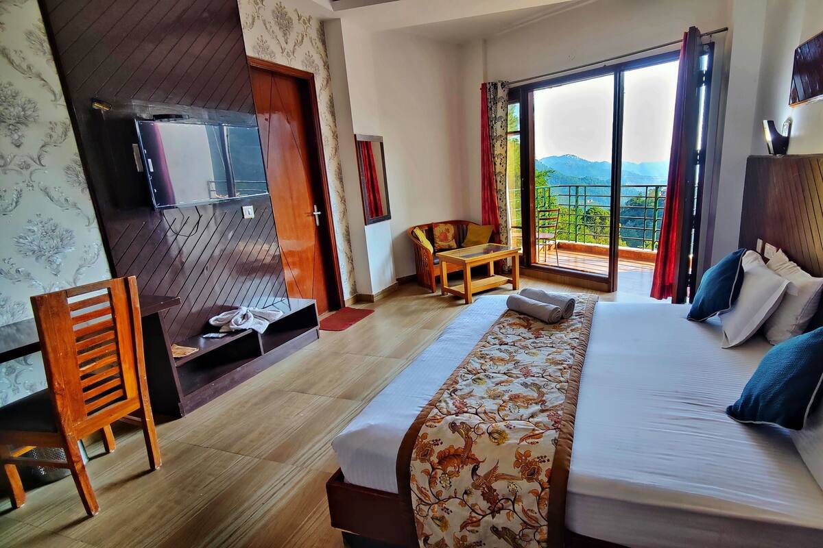 A hotel room with a bed, a chair, a small table, and a view of the mountains through the window.