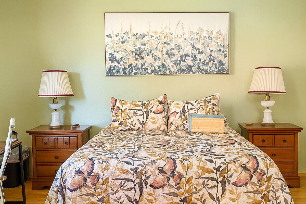 A bedroom with a floral bedspread, two bedside tables with lamps, a wooden nightstand, and a painting on the wall.