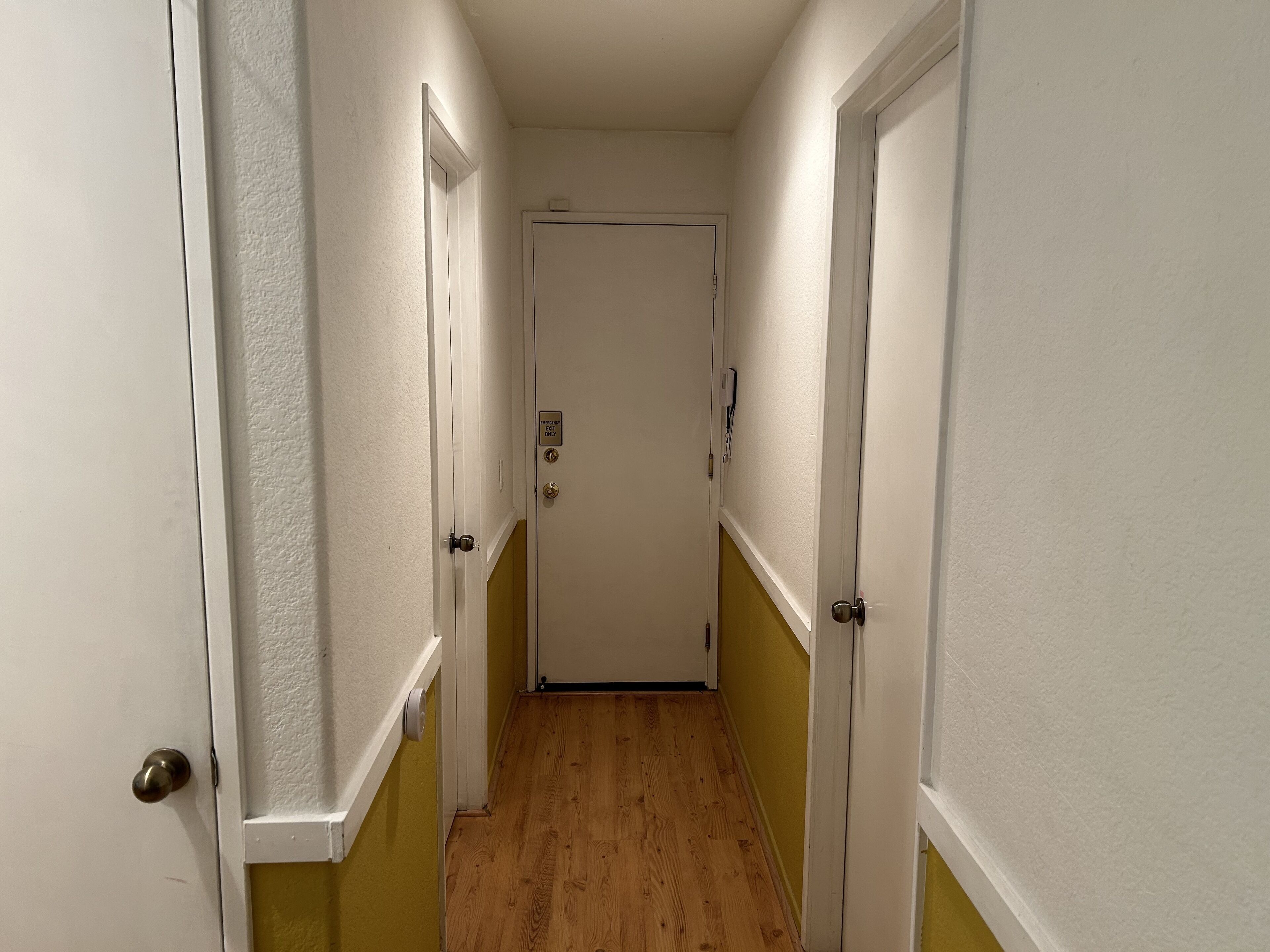 A narrow hallway with white walls and wooden flooring, leading to a door with an emergency exit sign.
