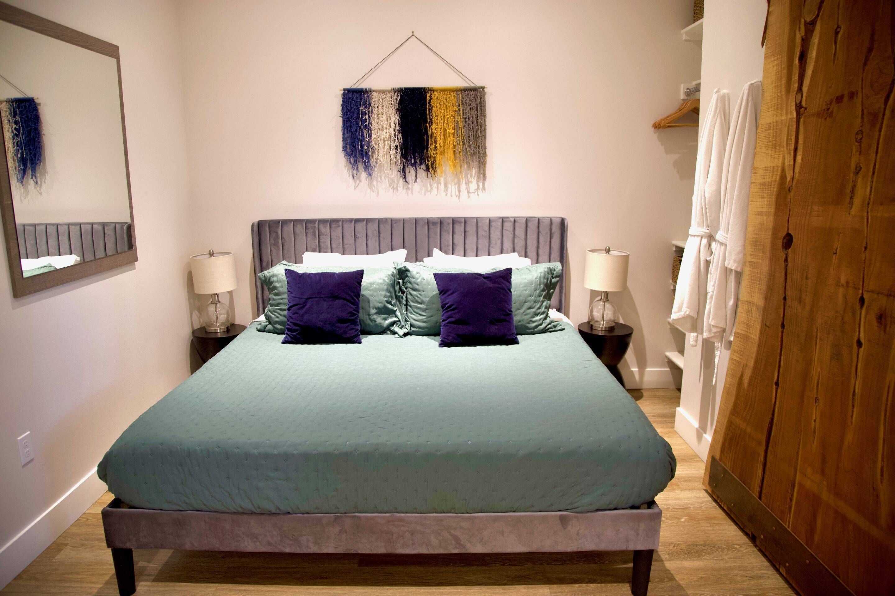 A neatly arranged bedroom with a large bed, two bedside tables with lamps, a hanging abstract art piece, and a wooden wardrobe.
