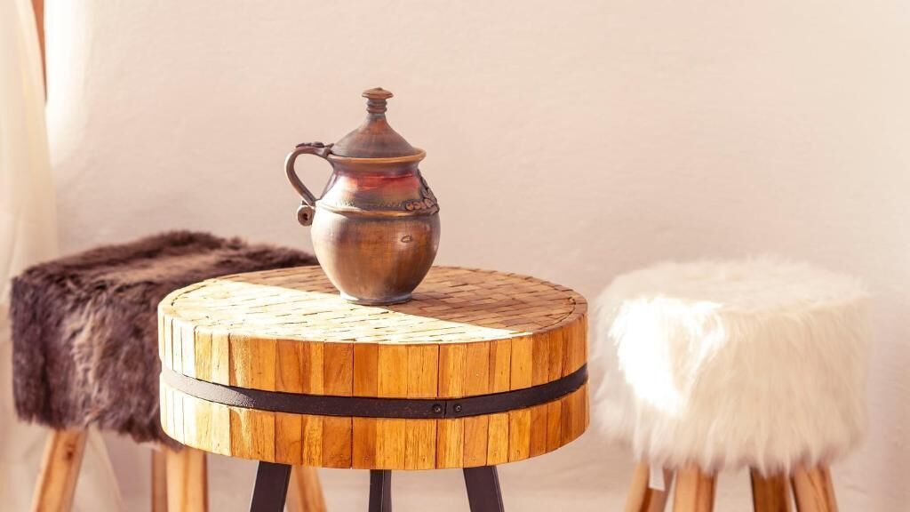 A round wooden table with a metal jug on top, flanked by a fur-covered stool and a white fur-covered stool.