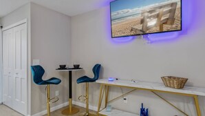 Ett rum med en tv-skärm som visar en strandscen, två blå stolar, ett litet runt bord och ett gyllene sidobord med en korg.
