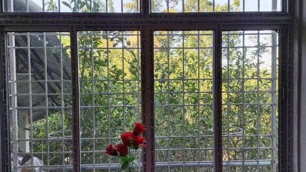 A window with metal bars and a vase with red flowers on a checkered table. - Homocation Changghar Resort (Kaliabar)
