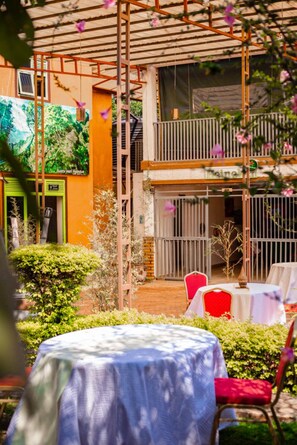 An outdoor patio area with tables and chairs, a wooden pergola, and a view of a building with a green door and a sign that reads "Heineken".