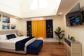 A bedroom with a bed, a bench, a nightstand, a lamp, a potted plant, and a TV mounted on the wall.