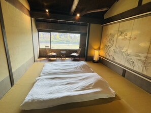 Ein Zimmer mit Tatami-Boden, einem Bett mit weißer Bettwäsche, einem Sessel, einem kleinen Tisch, einer Lampe und einem großflächigen Wandbild.