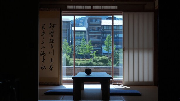A traditional Japanese room with tatami flooring, a low table with a bowl, and a sliding door with a view of buildings and greenery. - This is a single building rental of a Kyomachiya that has been in existence since the Meiji Era (1868-1912). The window overlooks the Kamo River and the town of Gion, making it a prime location to enjoy the typical Kyoto scenery. (Kyoto)
