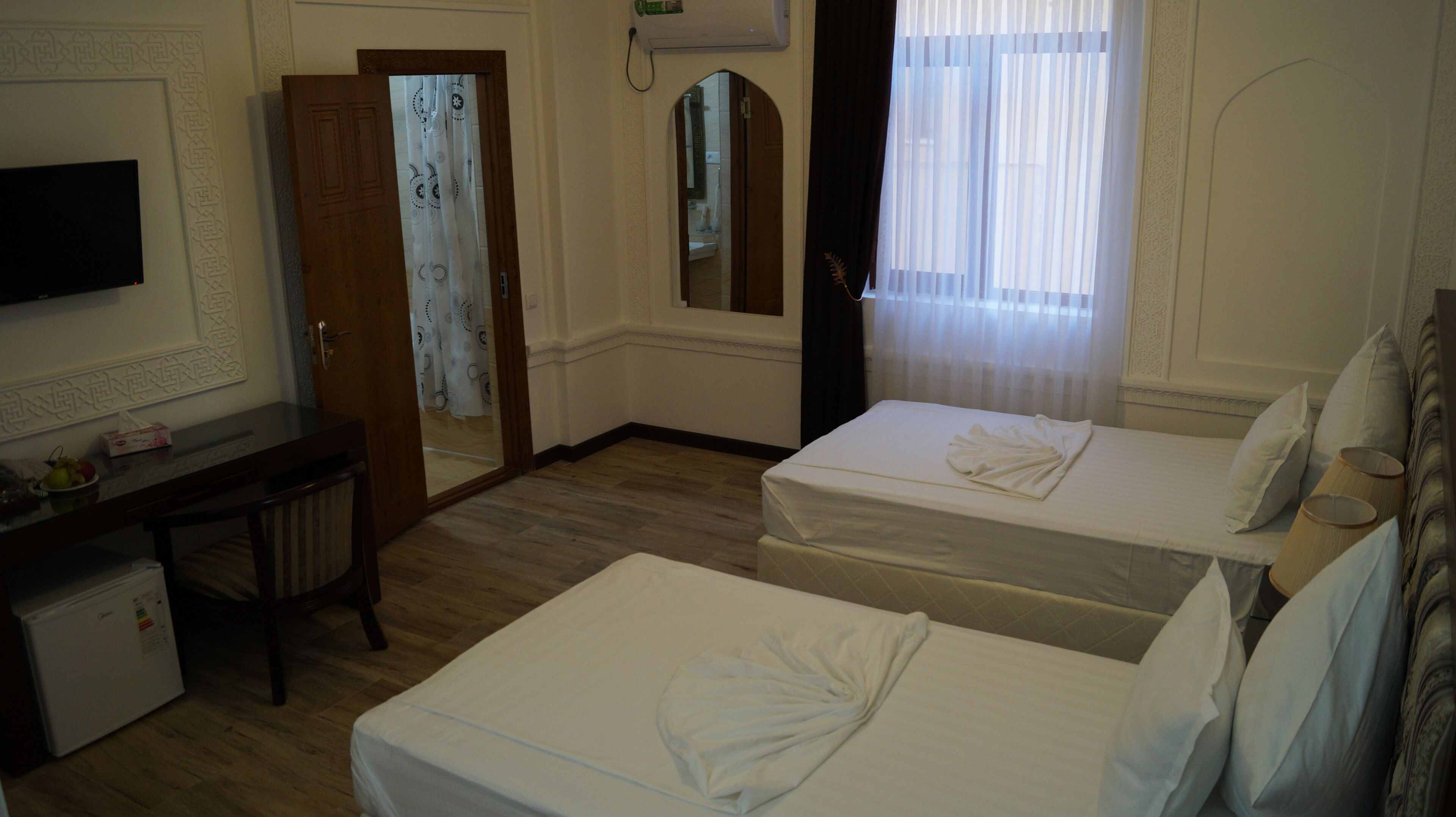 Une chambre d’hôtel avec deux lits, un bureau, une télévision et une salle de bain visible à travers une porte en arc de cercle.