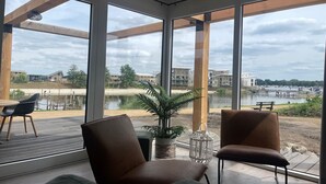 A modern room with large windows, a wooden deck, and a view of a lake and buildings. - Luxury Lodge with Beach View (Well)