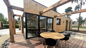 A wooden deck with a round table and four chairs, a glass door leading to an interior space, and a wooden exterior wall. - Luxury Lodge with Beach View (Well)