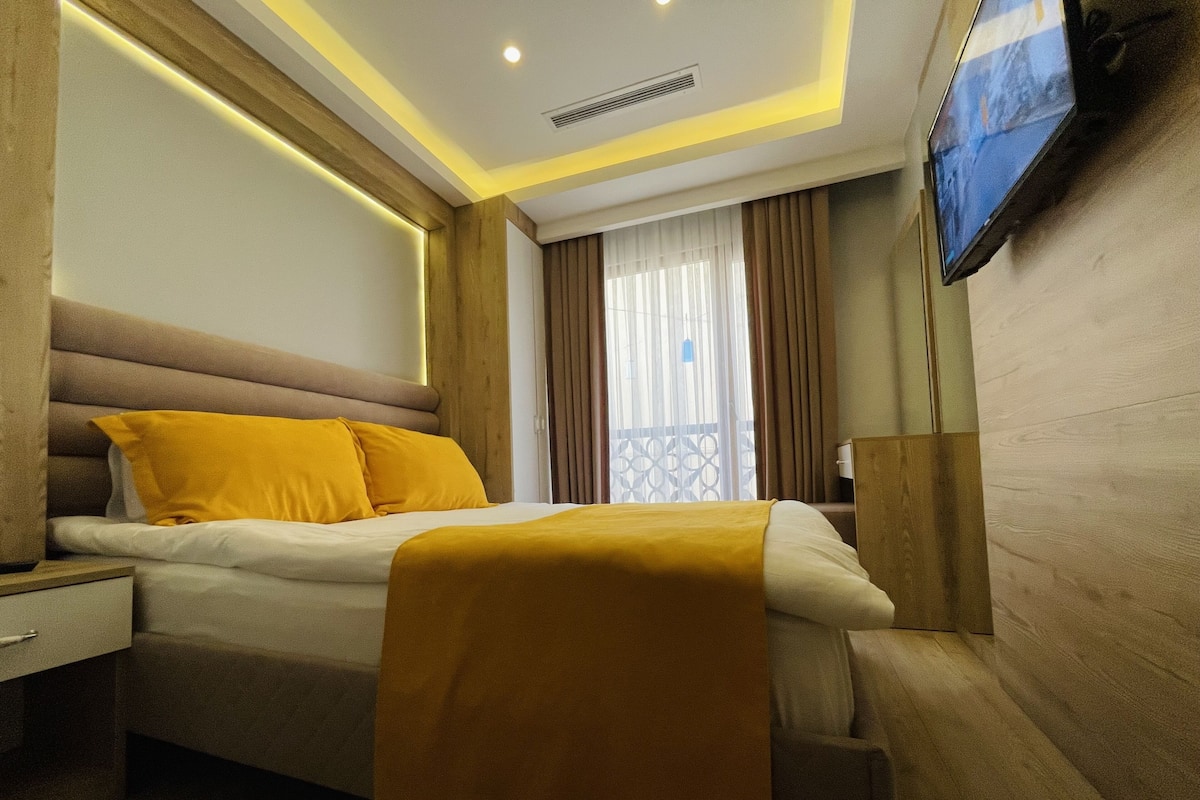 A modern hotel room with a large bed, a flat-screen TV mounted on the wall, and a window with curtains.