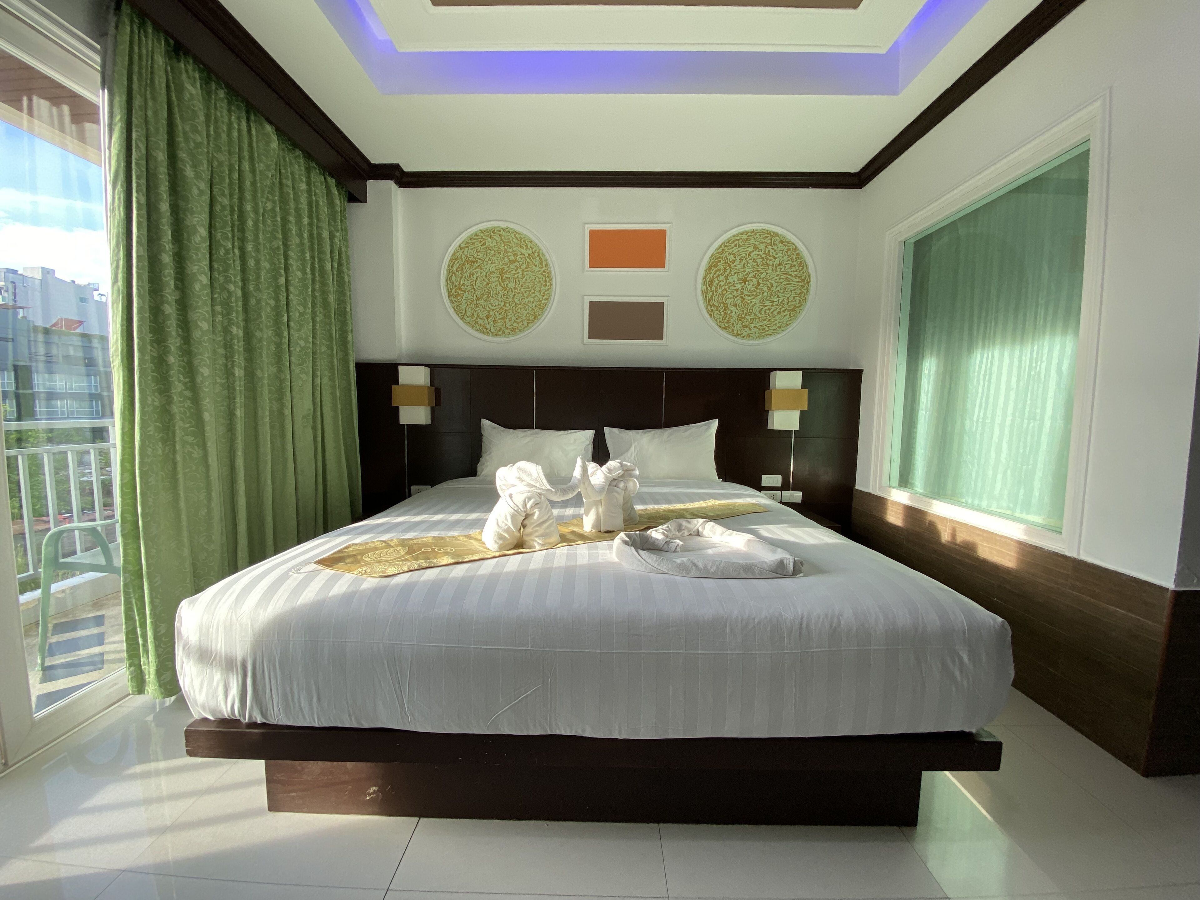 A large bed with a dark headboard, two wall sconces, and a view of the city through a large window with green curtains.