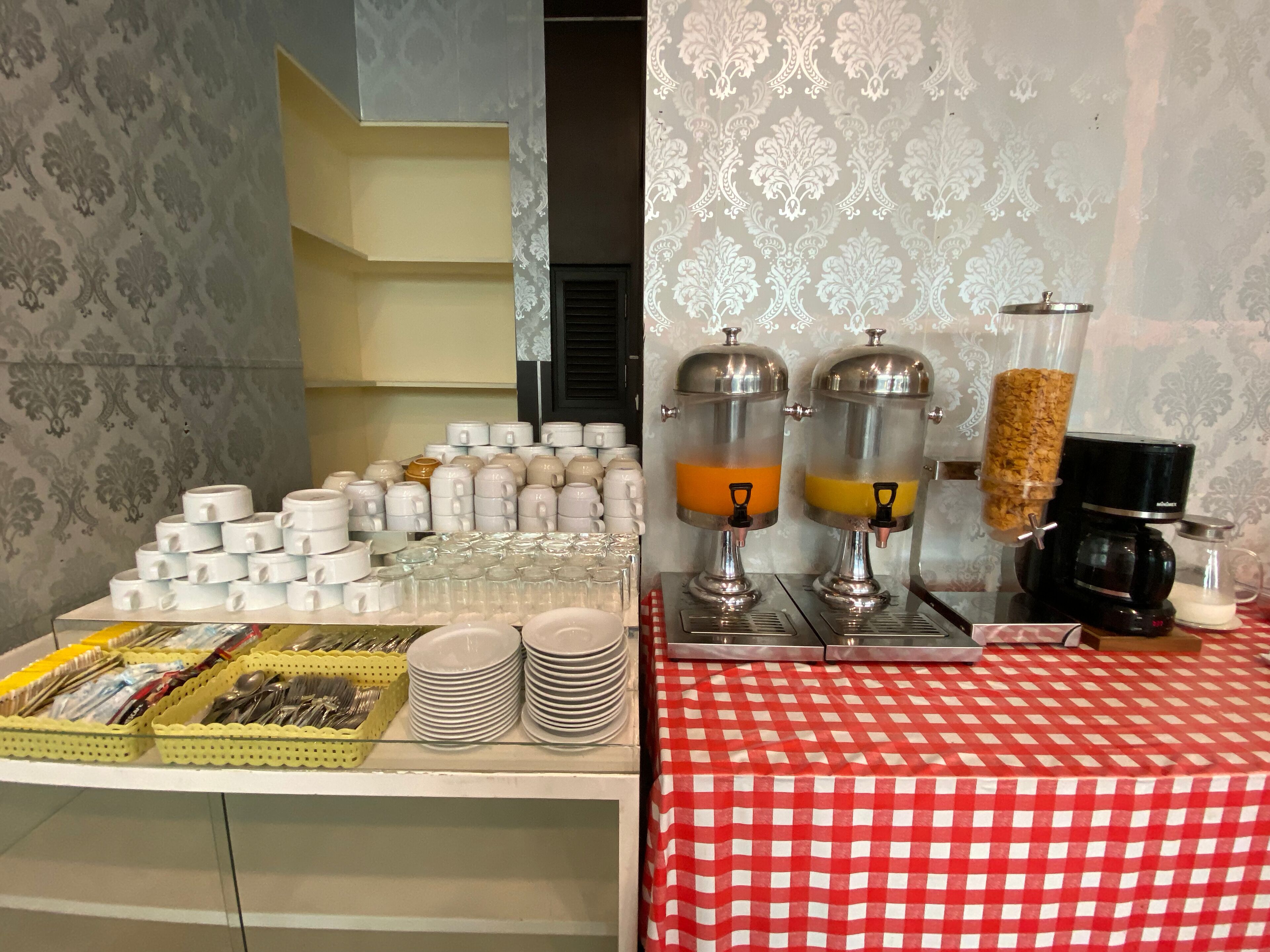 En morgenmadsbuffet med forskellige kopper, tallerkener og bestik, samt to drikkevareautomater med orange og gule drikke.