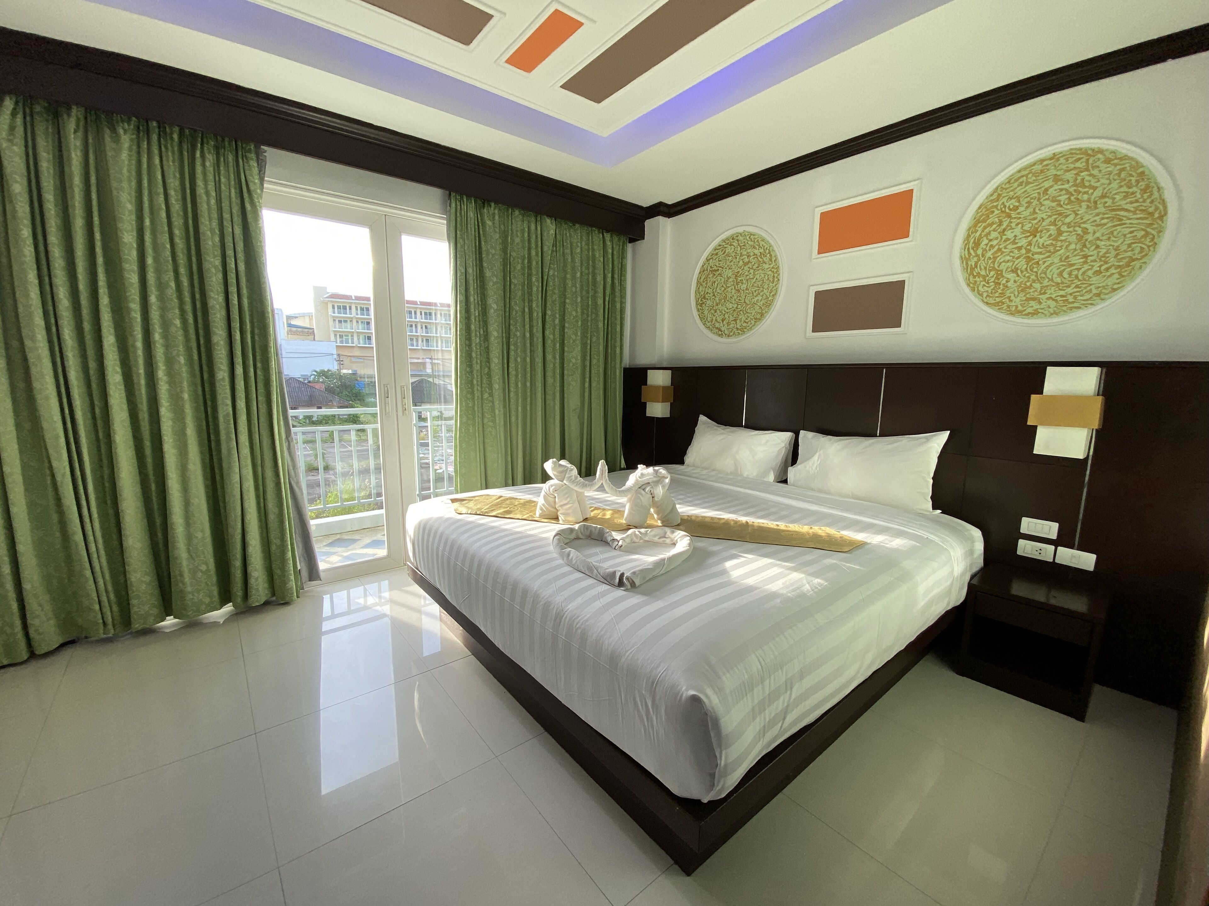 A hotel room with a large bed, green curtains, a view of buildings, and a ceiling with geometric patterns.