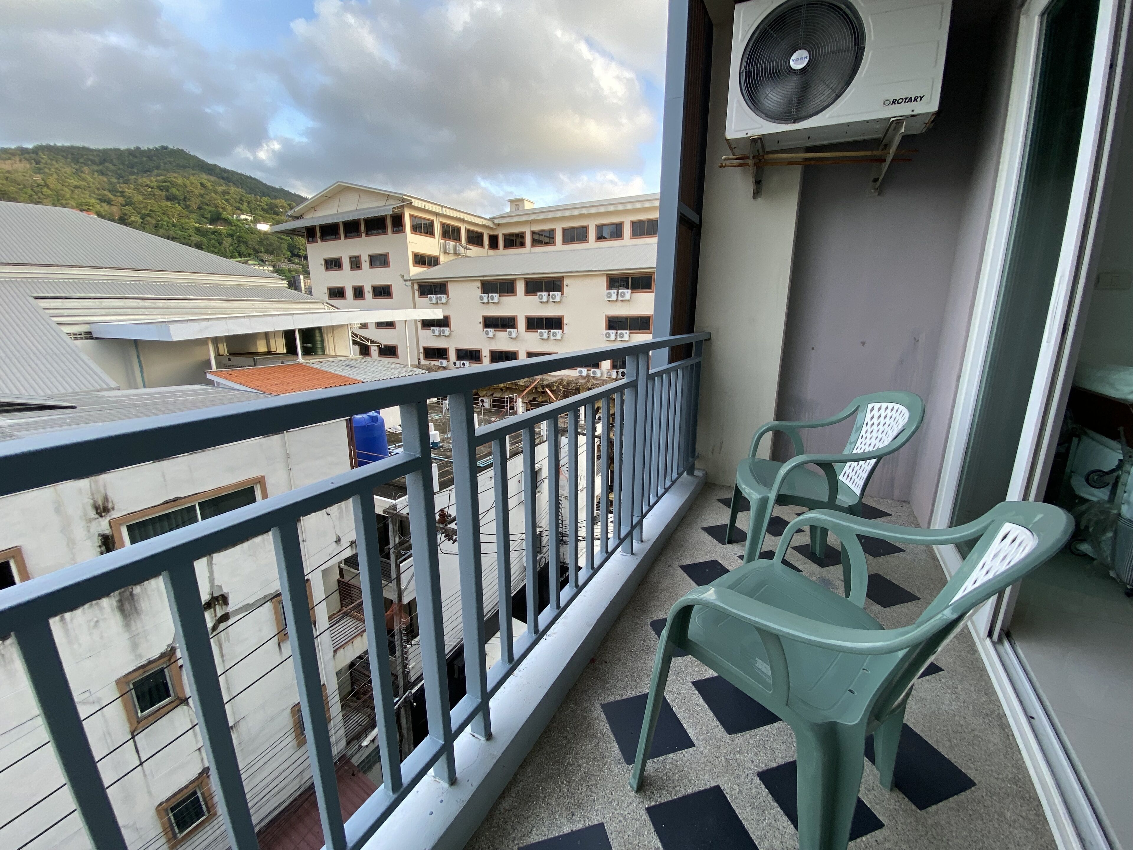 Een balkon met uitzicht op gebouwen, een ventilator en twee groene plastic stoelen.