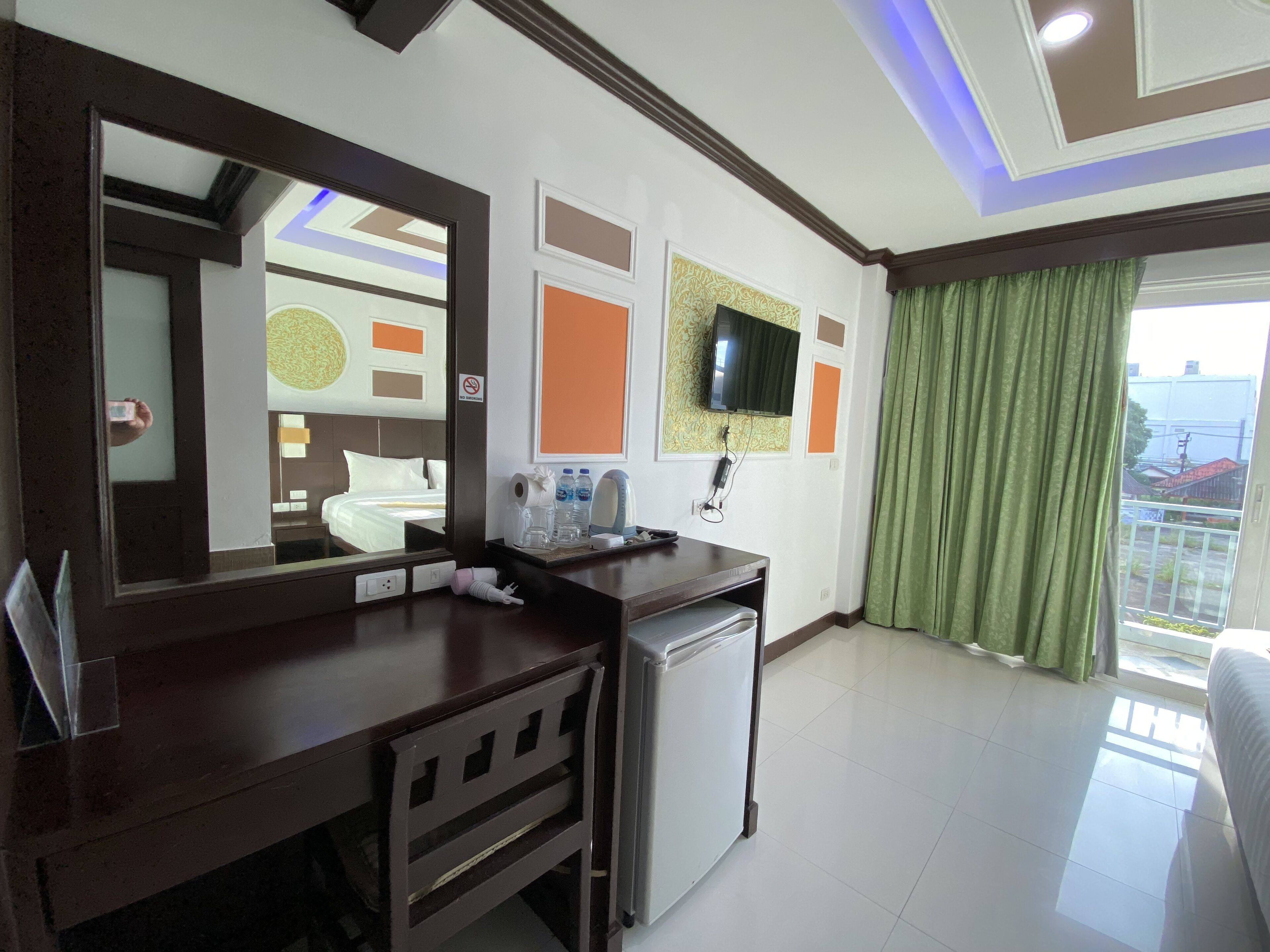 A hotel room with a bed, a desk with a mirror, a small refrigerator, a television mounted on the wall, and a balcony with green curtains.