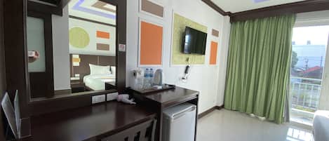 Habitación de hotel con cama, escritorio con espejo, refrigerador pequeño, televisor montado en la pared y balcón con cortinas verdes.