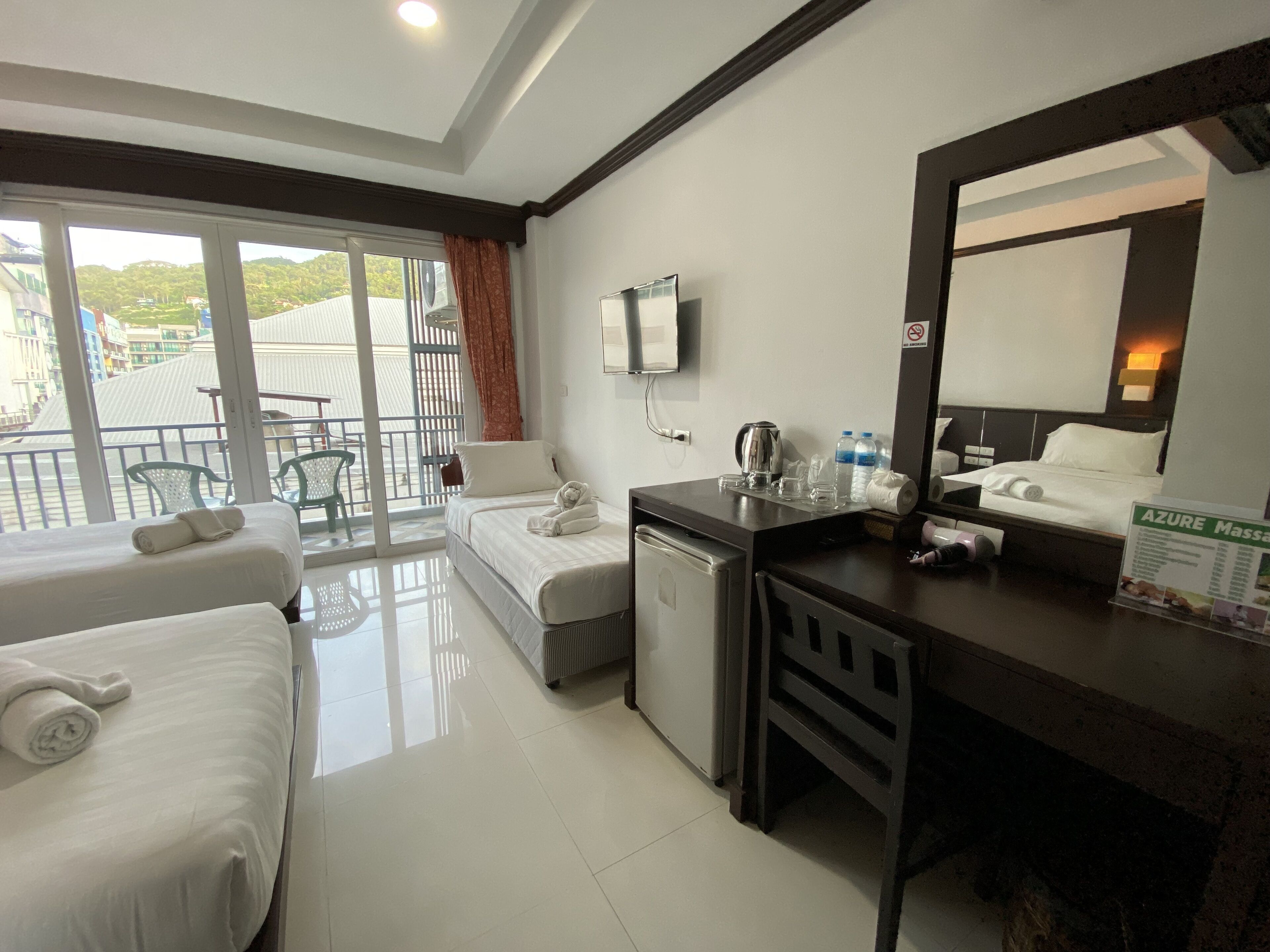 A hotel room with two beds, a desk, a mirror, a TV, and a balcony with a view of buildings.