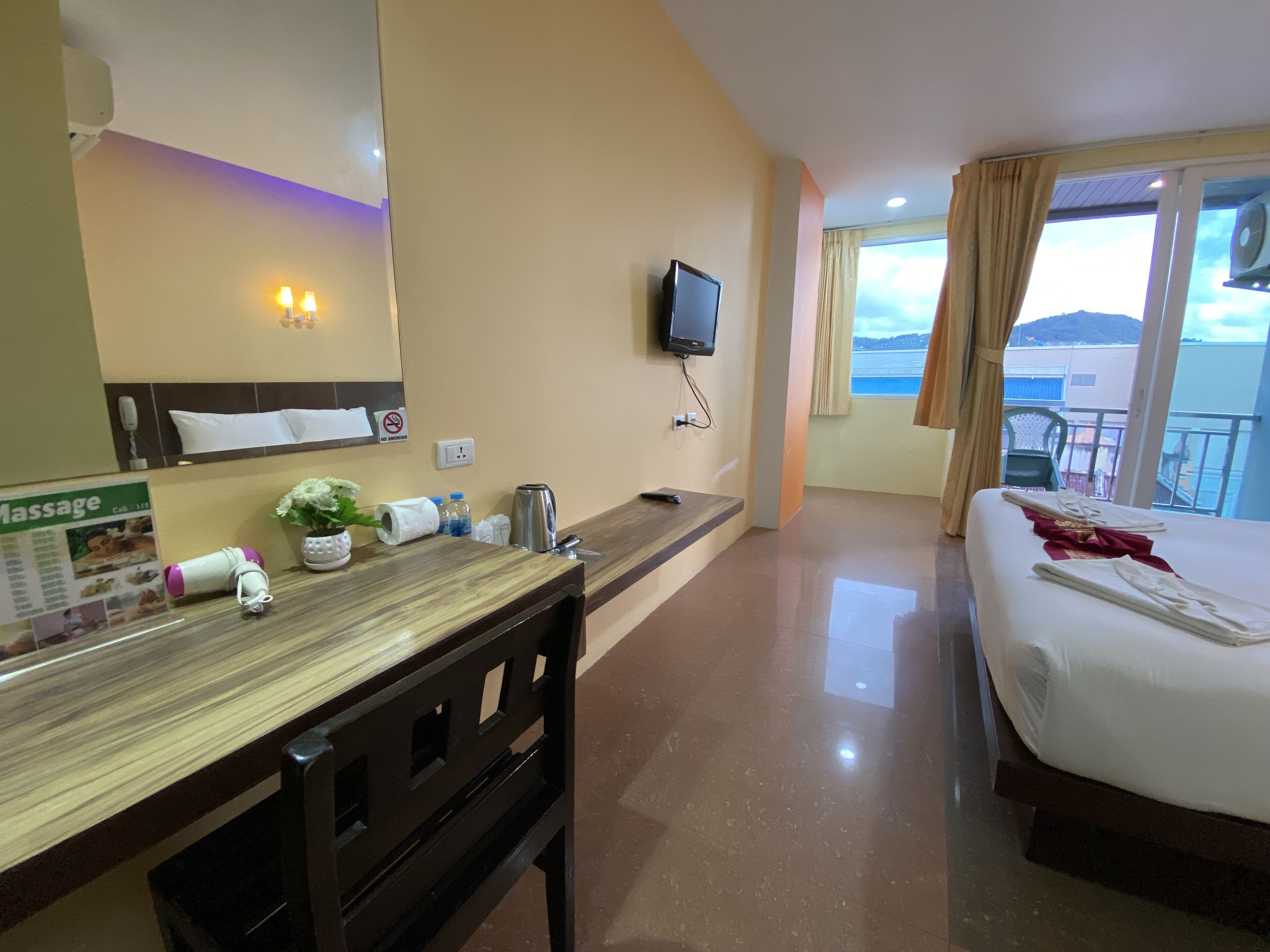 A hotel room with a bed, a desk with a chair, a TV, a massage brochure, and a view of the sea.