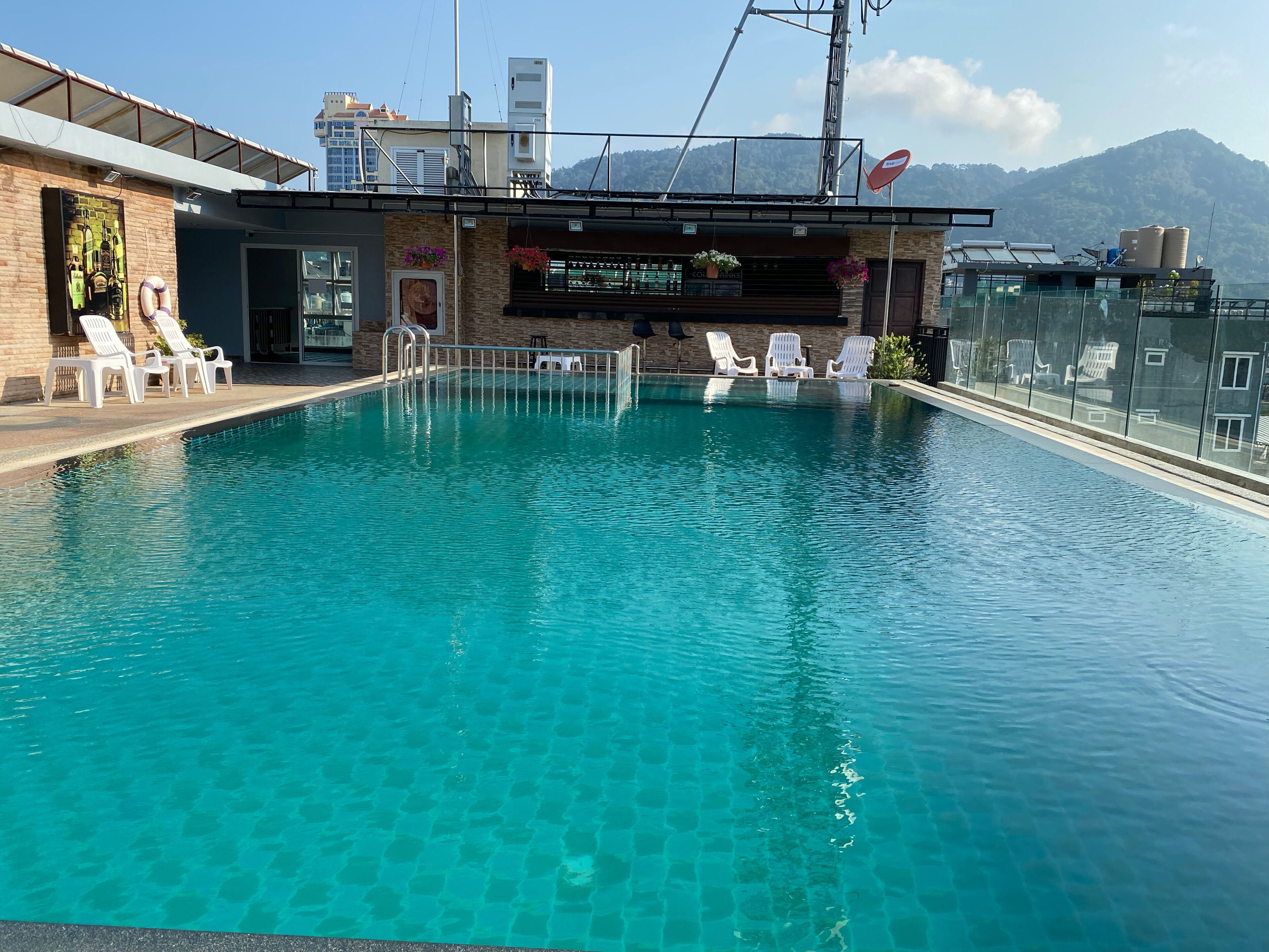 A clear blue pool with white lounge chairs, a brick building with a balcony, and a mountain backdrop.